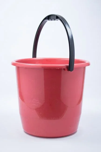 Dark plastic bucket half-filled with grass-infused water and a floating white Bti mosquito dunk in a backyard garden