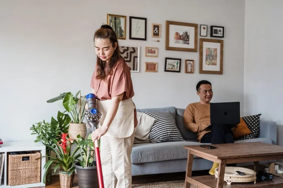 Person vacuuming a living room carpet and along the baseboards with an upright vacuum in daylight