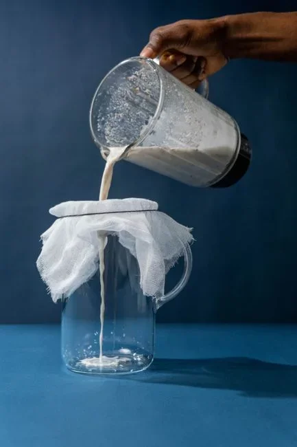 Hands pouring cold brew concentrate through a fine mesh strainer lined with cheesecloth into a second glass jar on a kitchen counter