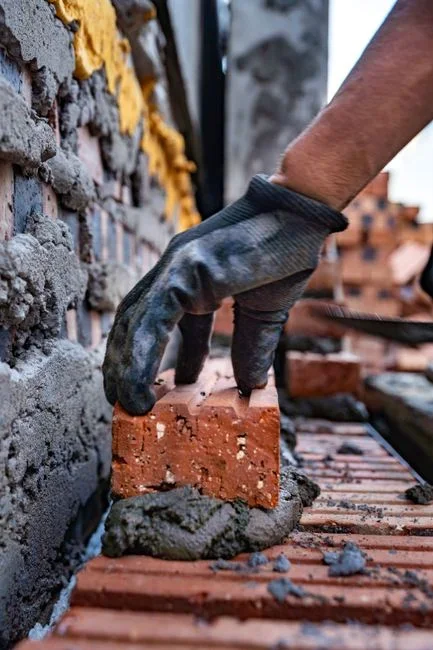 Gloved hands using a narrow hand chisel and mallet to rake crumbling mortar out of a single brick joint