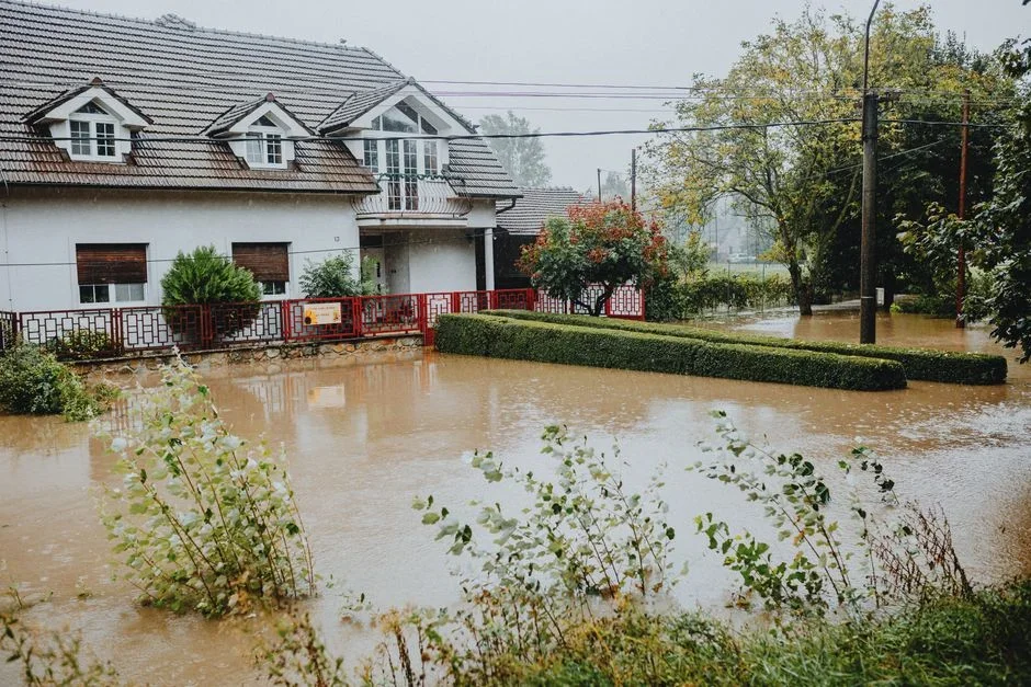 Muddy brown floodwater surrounding a rural well head and pump house after heavy rain, debris caught against the casing