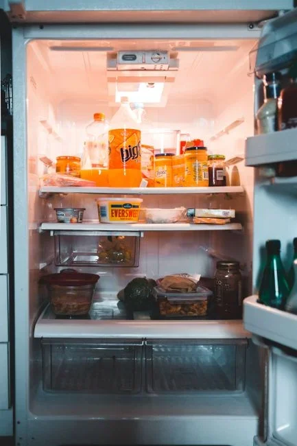 An appliance thermometer standing in a glass of water on a refrigerator shelf beside milk and vegetables