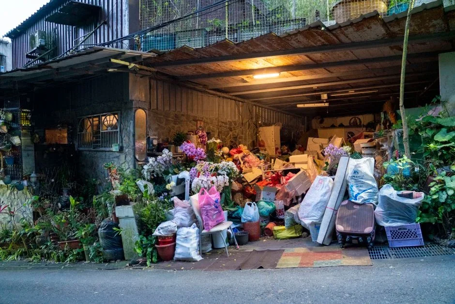 An old beige refrigerator plugged in against the wall of a cluttered suburban garage next to storage bins and a lawn mower