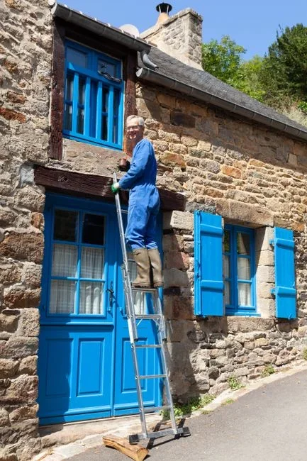 Man standing on an extension ladder scooping wet leaves out of a single-story house gutter
