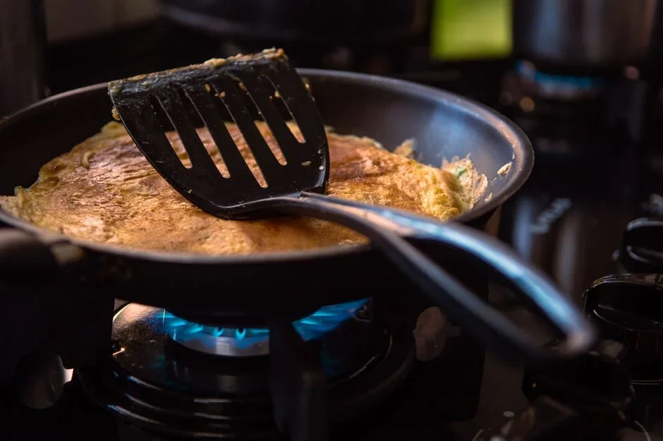 A metal pan lid being slid across a smoking skillet on a stovetop burner to smother flames, with the burner control knob turned to off