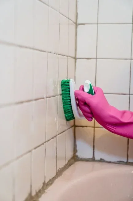 Stiff-bristled scrub brush, bucket of hot soapy water and yellow rubber gloves on a bathroom floor beside tile