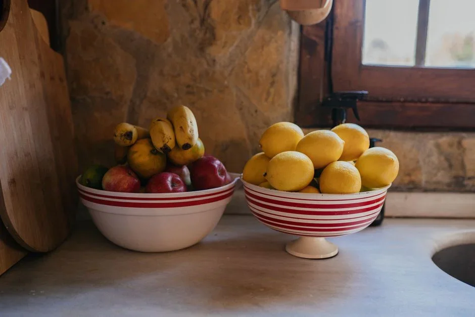 Bunch of yellow bananas sitting in a wooden fruit bowl next to red apples and tomatoes on a kitchen counter in warm daylight
