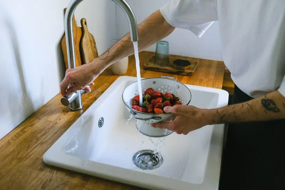 Fresh strawberries in a metal colander being rinsed under running tap water at a kitchen sink
