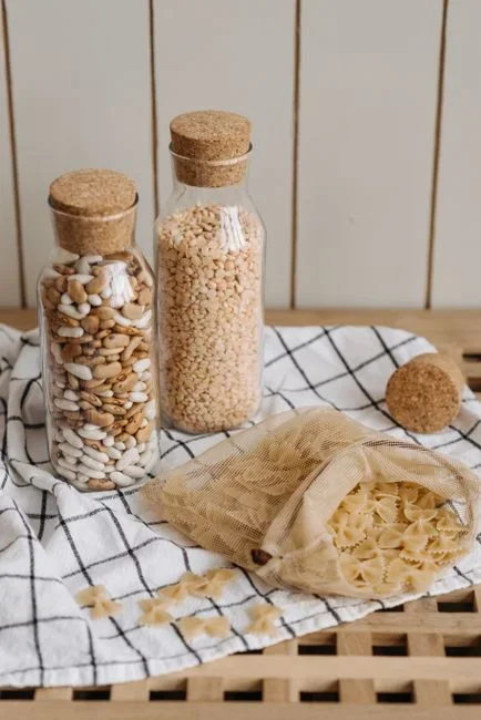 Row of screw-top glass jars holding rice flour and pasta on a pantry shelf next to a torn paper flour bag with spilled grain