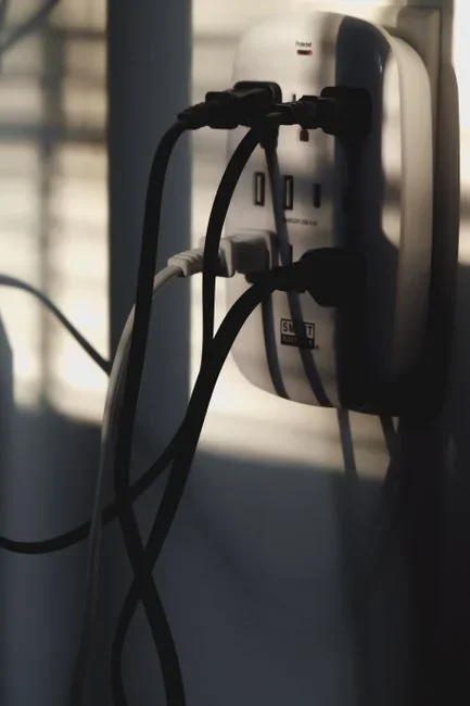 Close-up of a cluttered power strip behind a TV stand with a cable box, sound bar and tangled cords, small standby LEDs glowing in a dim living room