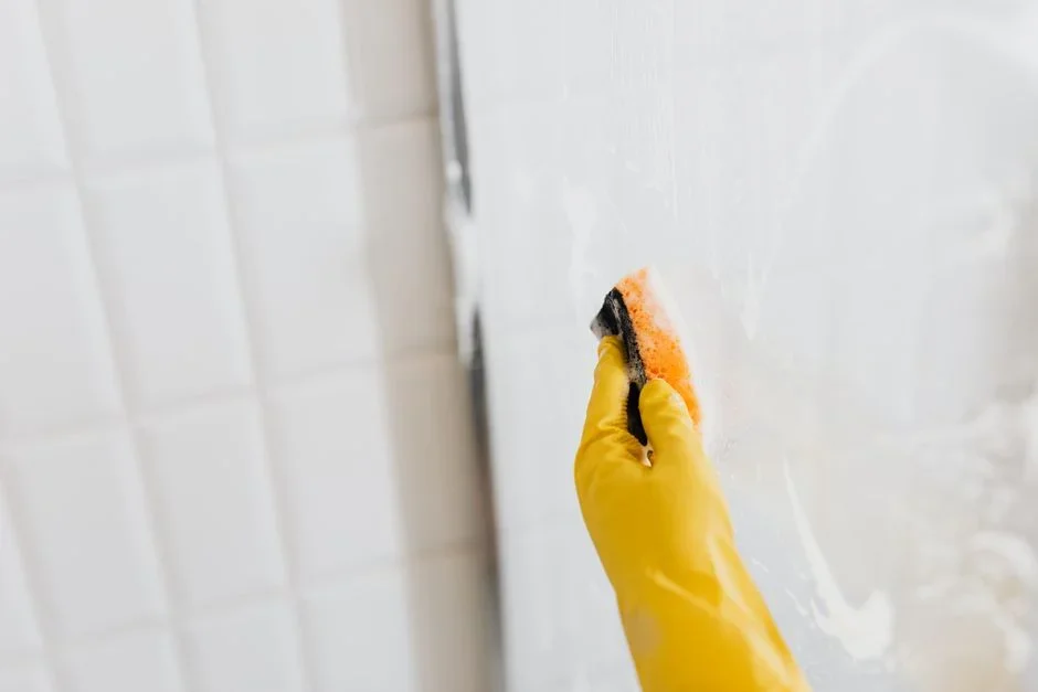 Person using a rubber squeegee to pull water down a wet glass shower door in a bright bathroom