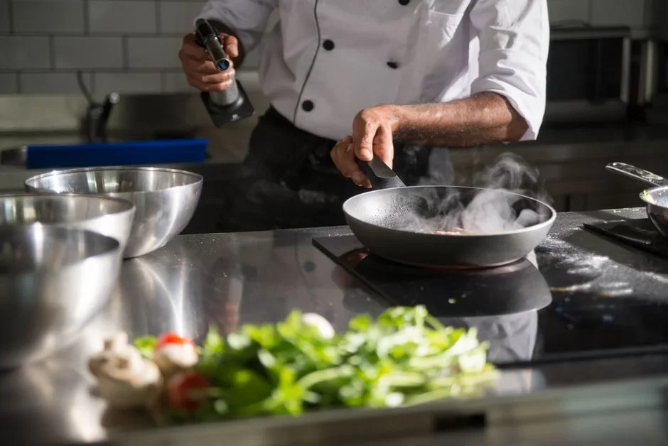 A hand reaching up to pull the battery out of a ceiling smoke alarm while smoke rises from a pan on the stove behind