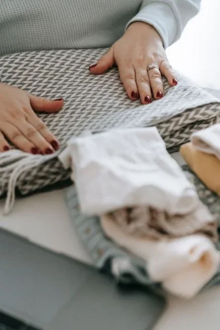 Hands folding a clean wool sweater beside an open washing machine and a stack of folded knitwear on a laundry room counter